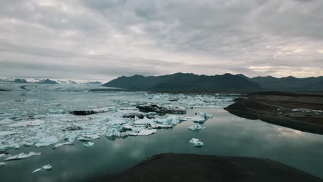 Melting-glacier-due-to-global-warming-with-icebergs-drifting-on-its-surface,-Mountains-under-an-overcast-sky-in-the-background,-Iceland,-Slow-motion-drone-shot