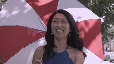 happy asian woman strolls down a city street carrying a red and white parasol on a sunny day