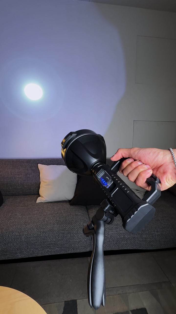Close-up of a hand holding a rechargeable LED flashlight in a modern living room, casting a bright circular beam on the wall beside a gray sofa and cushions.