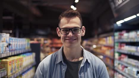 primer plano del apuesto joven caucásico con gafas haciendo compras en el supermercado y sonriendo alegremente a la cámara. retrato