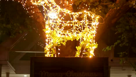 Leuchtendes-Weißes-Rentier-Auf-Dem-Frohe-Weihnachten-Podium-Mit-Beleuchtetem-Baum-Im-Hintergrund