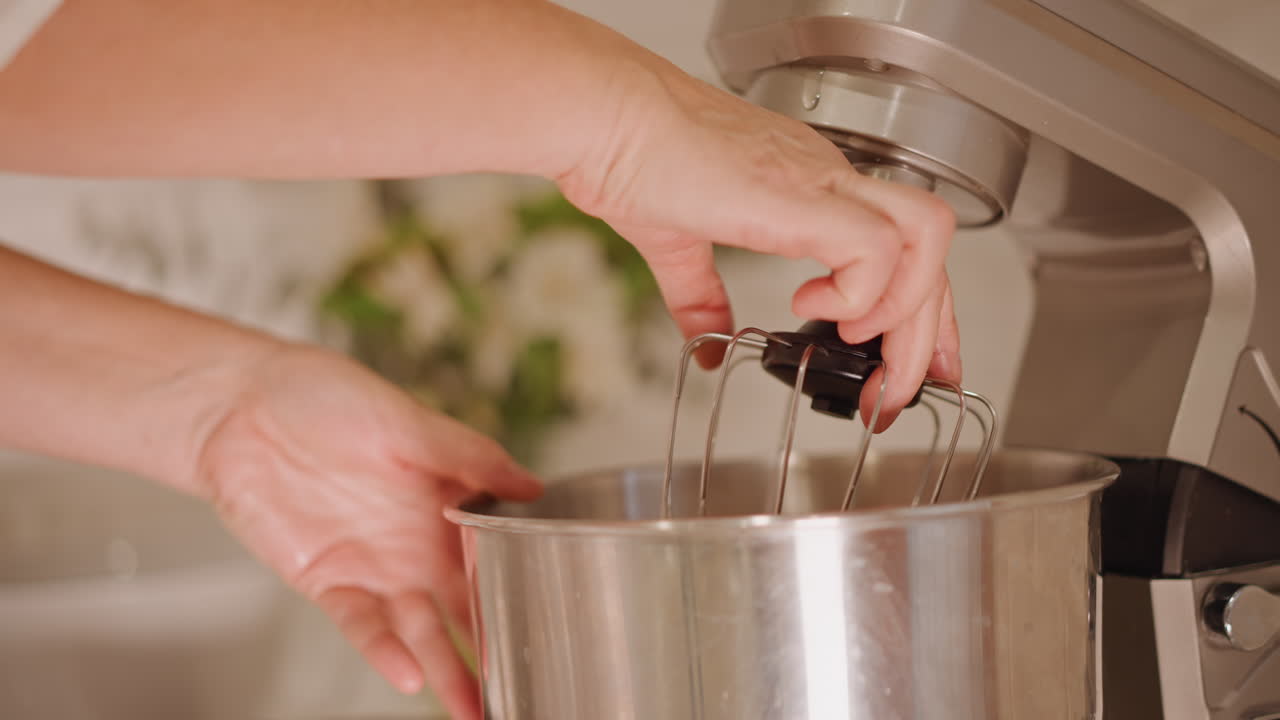 Mani che attaccano la frusta del mixer alla ciotola, una casalinga assembla l'accessorio della planetaria per il ripieno della torta di zucca in una cucina accogliente, primi piani di ciotola in acciaio inossidabile, frusta, mani, luce naturale calda, sfocato.