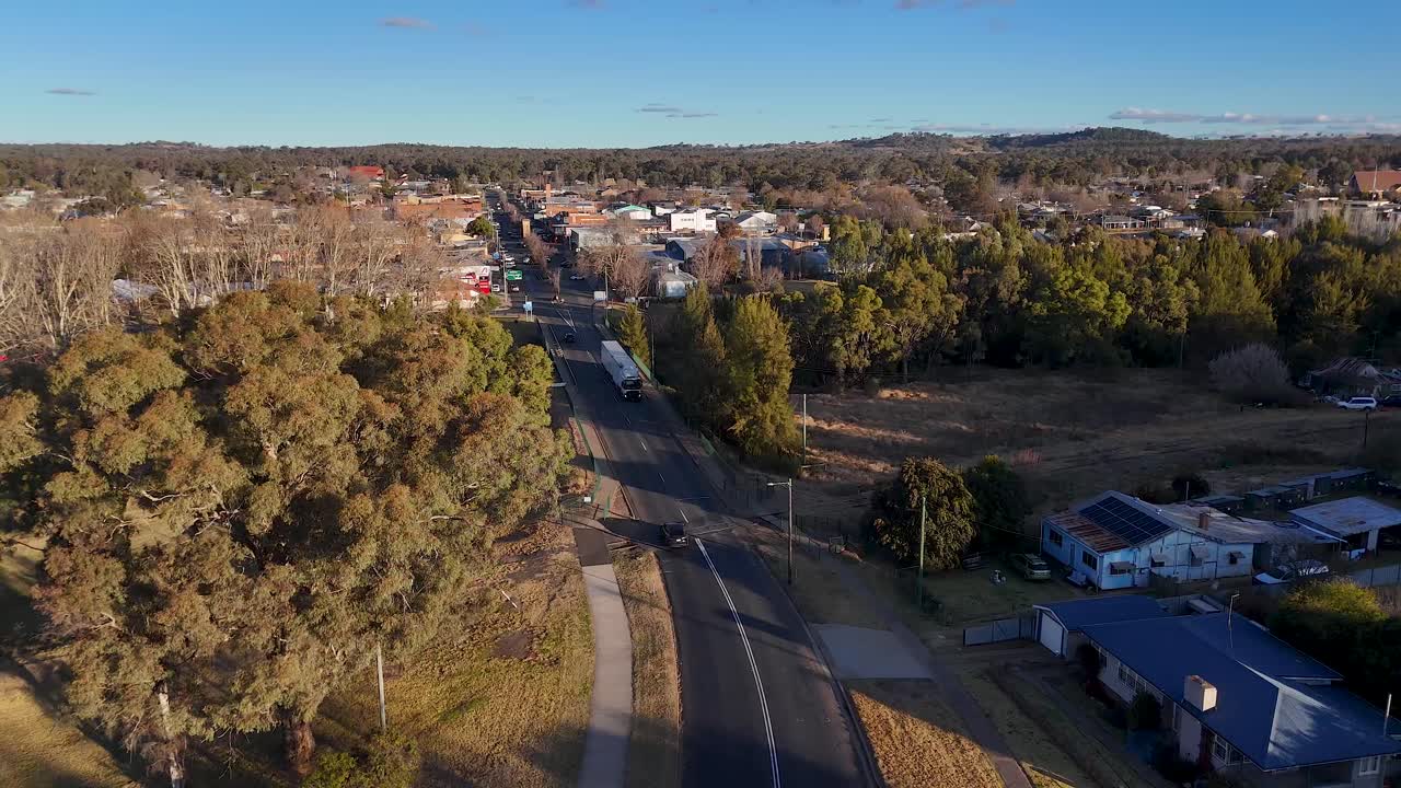 Aerial tracking shot of a car driving along a tree-lined street in a quiet residential area during golden hour, revealing a small town center