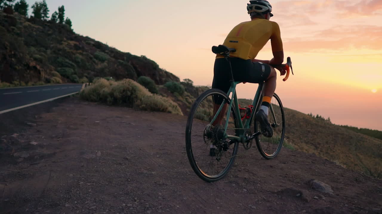 Sporting yellow t-shirt and gear, athlete rests on bike at mountain's zenith, savoring mountain vista and sunset after training