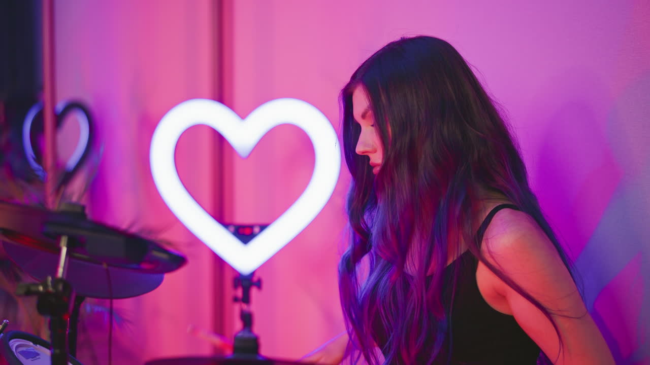 Side view of lady with long flowing hair covering face while playing drum beside heart-shaped ring light in vibrant studio with pink and purple lighting casting soft shadows and dreamy ambiance
