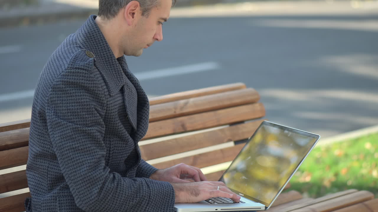 Man in a grey coat working on her laptop on a bench in the park