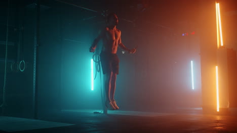 Muscular-man-with-bare-torso-is-jumping-using-a-skipping-rope.-Male-boxer-with-skipping-rope.-Fighter-doing-jump-rope-exercises-in-colorful-light-with-smoke-in-slow-motion.