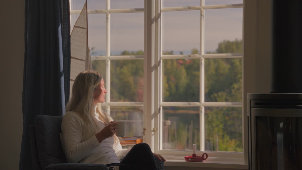 Caucasian blonde woman reading a book in a comfortable chair by a window, then sipping a warm beverage while looking at a peaceful lake and forest in soft natural light.