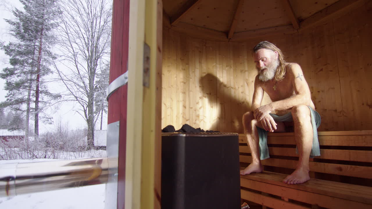 Long-haired scruffy man unwinds in hot steamy wooden sauna as it snows outside