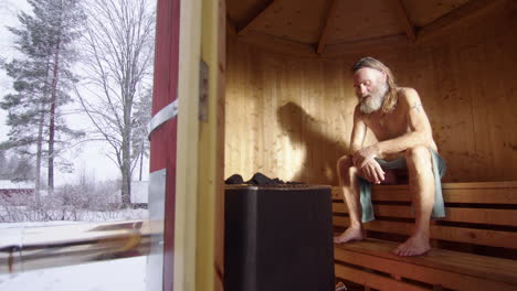 long-haired scruffy man unwinds in hot steamy wooden sauna as it snows outside