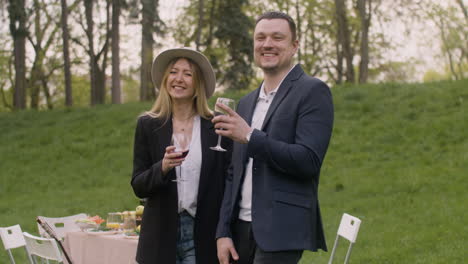 middle aged man and woman laughing and looking at camera while toasting with wine glasses in the park