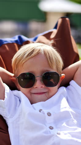 funny blond little boy wearing big sunglasses spends time outdoors. cute kid folded his arms behind the head lying in a comfortable chair. vertical video