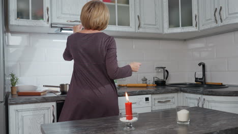 back view of lady dancing in modern kitchen, wearing plaid clothing, illuminated by candlelight, white cabinetry, dark countertops, organized space with rolling pin, kettle and sink