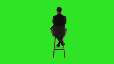 rear view studio shot of man sitting on stool facing away from camera against green screen