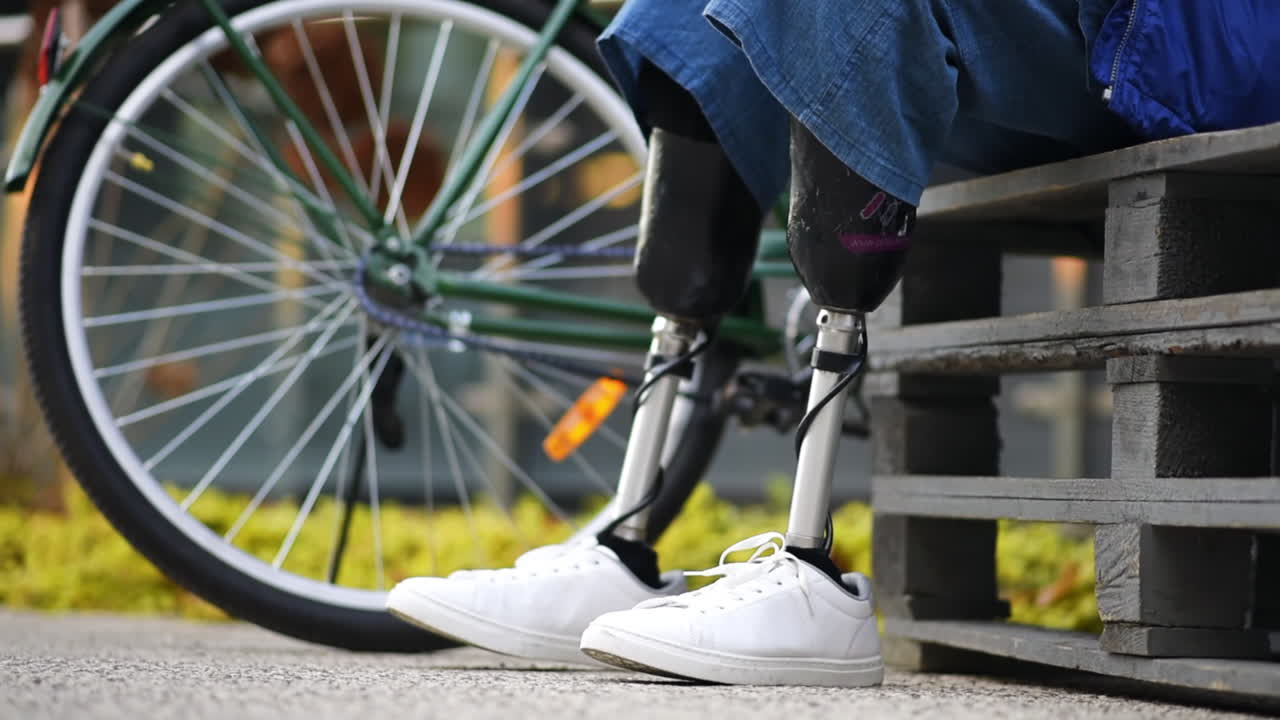 Slow motion view of a man with prosthetic legs. Sitting down on a bench, bicycle nearby