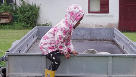 Athletic-little-African-girl-in-pink-raincoat-with-hoodie-playing-outdoor-climbs-off-tractor-trolley-in-countryside-farm,-handheld,-rainy-day