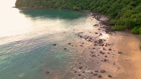 Aerial-drone-view-of-beach-with-trees-and-rocks,-clear-blue-waters-during-sunset