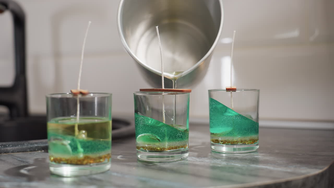 Hand view of mother in kitchen pouring melted sugar into second glass container containing green decorative substance with wick, crafting homemade project with precision on clean countertop surface