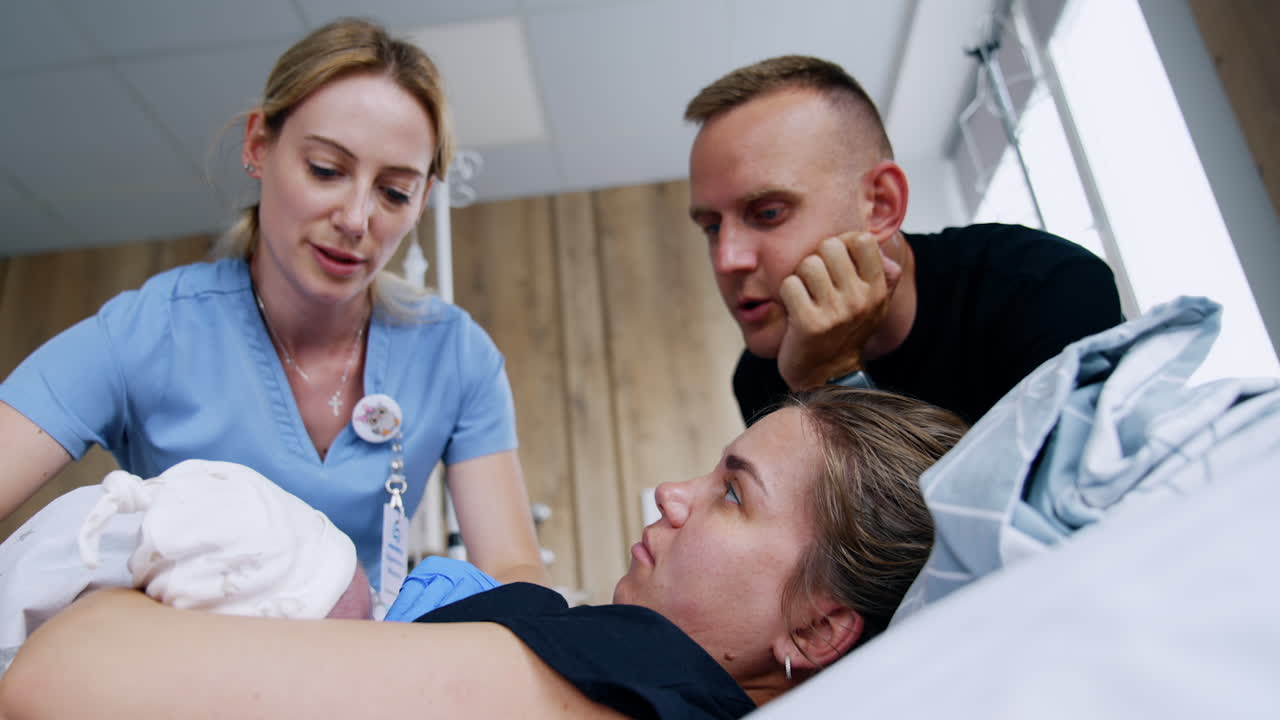 Female consultant on breastfeeding helping young mom to feed her newborn. Happy dad stands beside asking questions. Low angle view.