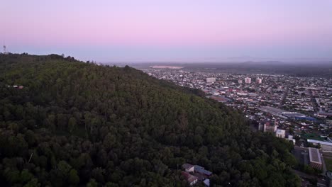 Fliegen-Sie-über-Den-Cerro-Ñielol-Und-Die-Stadt-Temuco-Mit-Einem-Violetten-Sonnenuntergang-Und-Dem-Vulkan-Laima-Im-Hintergrund