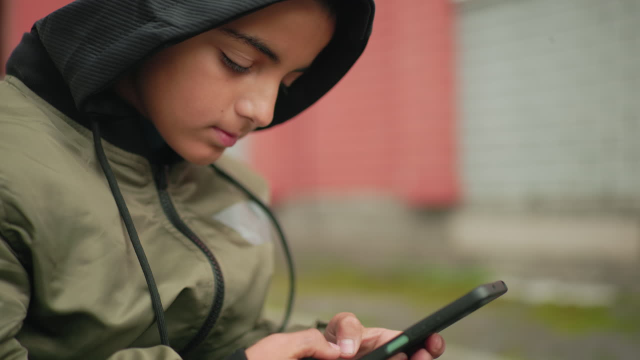Close up of kid in hoodie adjusting head slightly with eyes closed, showing thoughtful expression against blurred urban background with red and gray walls creating moody atmosphere