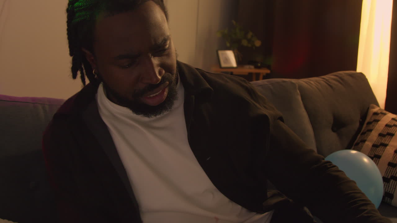 Man Sitting On Sofa At House Party Looking Through Half Empty Glasses And Bottles Of Alcohol
