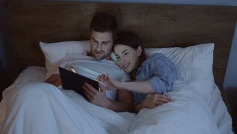 top view of smiling caucasian wife and husband lying in the bed using tablet under blanket late in the evening