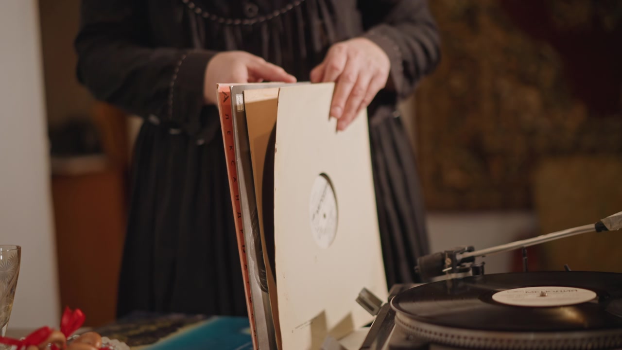 Žena vybírající vinyl ze stohu vedle gramofonu, opatrné ruce obracejí kartonové obaly a prohlížejí štítky, stůl s občerstvením a vintage dekorem vytváří útulnou sběratelskou atmosféru a hmatovou nostalgii.