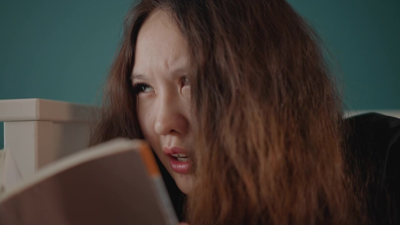 Close-up shows young woman with long wavy brown hair seated beside white bedpost in softly lit bedroom, reading book with downward gaze, parted lips, and thoughtful speaking reaction