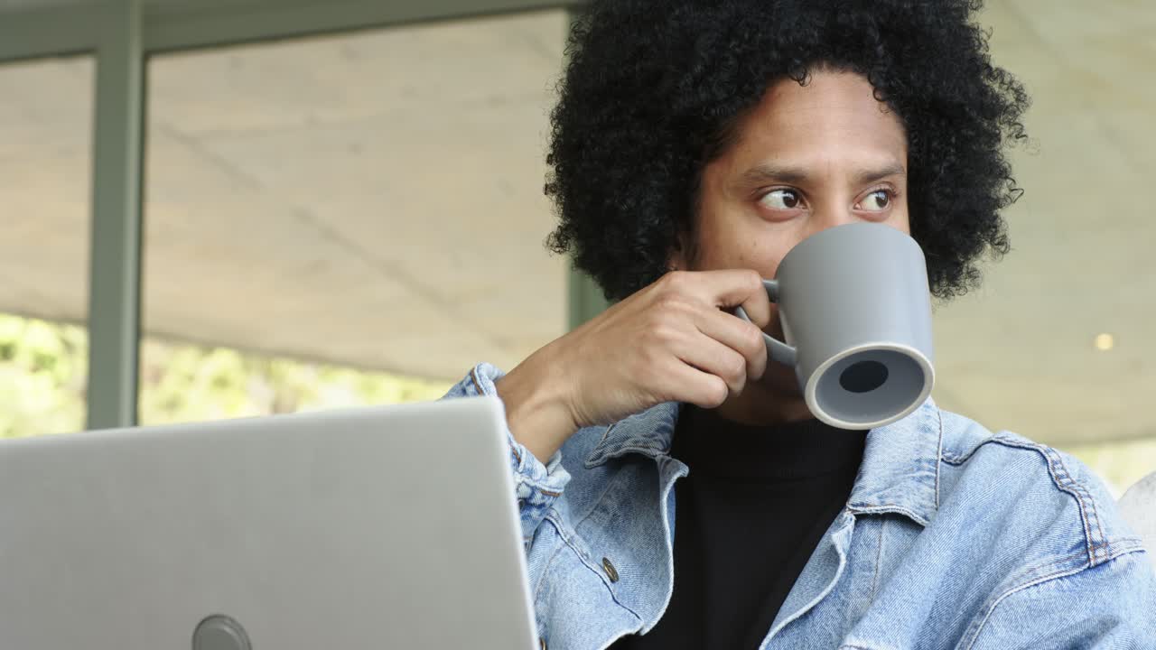 En Afroamerikansk mand søger kaffe, tager et krus ved sin laptop, nipper til det på verandaen, med plads til tekst.