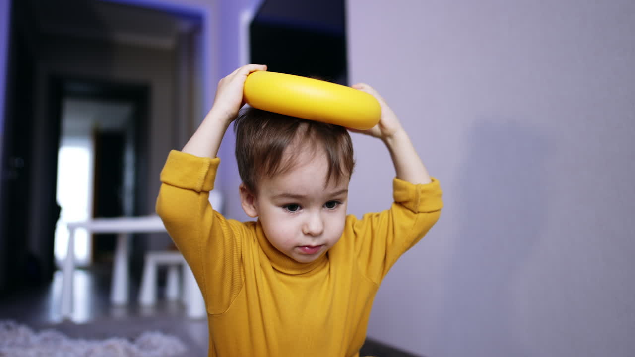 Beautiful brunet male kid in yellow sweater plays with toys. Toddler places ring from toy pyramid on his head and drops it. Close up.