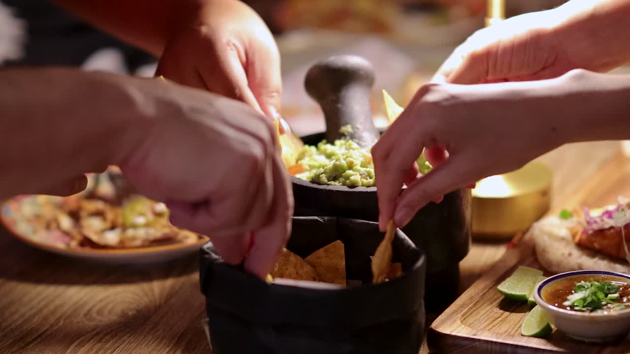 Multiple hands reach for guacamole with tortilla chips around a festive table in warm lighting, highlighting a lively social gathering in Bangkok