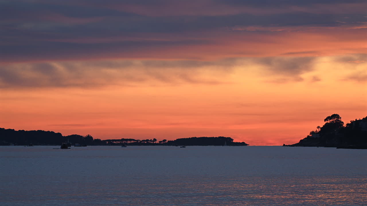 Wide shot of calm sea under a colorful sunset sky, with distant islands and boats silhouetted on the horizon