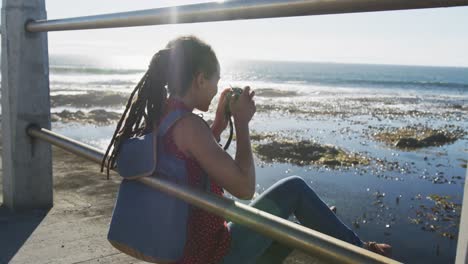 mujer afroamericana sentada y fotografiando en el paseo marítimo por el mar