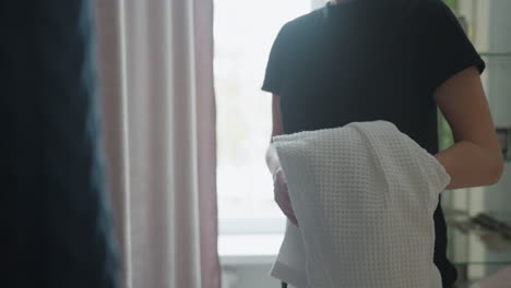 woman extends hand holding white textured robe toward another lady while making gentle gesture, captured in softly lit room with blurred background, showcasing kindness, care, and interaction