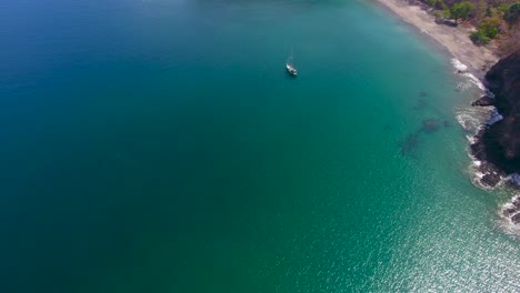 Aerial-video-flying-down-on-a-single-monohull-sailboat-anchored-in-an-empty-calm-bay-tilting-up-to-reveal-a-beautiful-sandy-beach-in-paradise