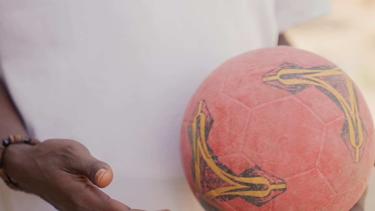 Mãos de treinador negro oferecendo bola remendada, troca em rua ensolarada destacando palma, pulseira de contas e detalhe do remendo, treinamento de alcance comunitário e gesto de passagem para prática juvenil.