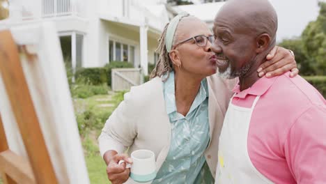 Happy-senior-african-american-couple-hugging-and-painting-on-wooden-easel-in-garden,-slow-motion