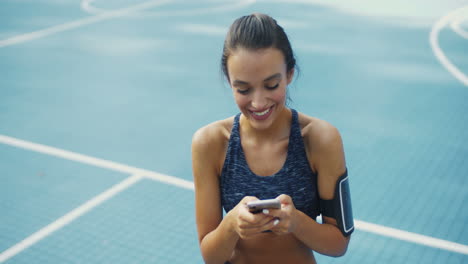 chica deportiva con airpods riéndose mientras envía un mensaje de texto en un teléfono inteligente en la cancha al aire libre en un día de verano 2