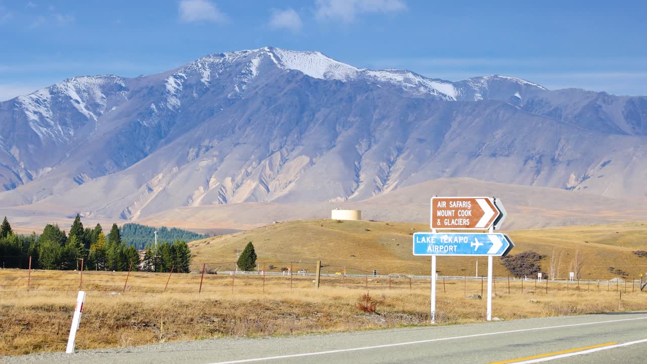 A silver SUV drives past a road sign on a rural highway with snow-capped mountains in the background, under bright daylight and clear skies