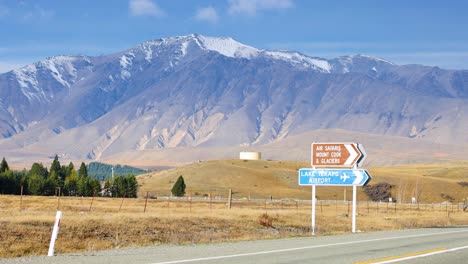 a silver suv drives past a road sign on a rural highway with snow-capped mountains in the background, under bright daylight and clear skies