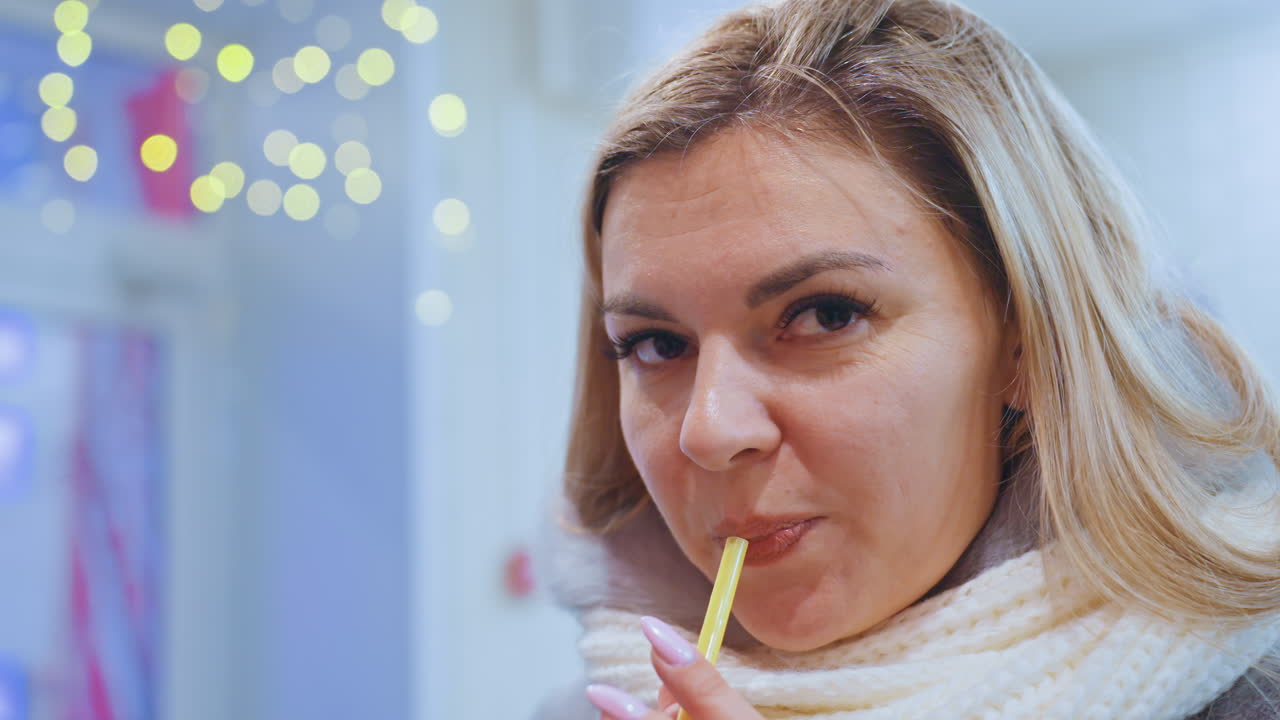 Kaukasisk kvinne nipper til kaffe med sugerør, iført tykt hvitt skjerf, står inne i en festlig butikk med glitrende bokeh-lys, nærbildeportrett som viser et avslappet smil, ærlig uttrykk.