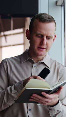 young man leaned on the window holding a telephone and a paper notebook. man turning pages in a book and comments something. vertical video