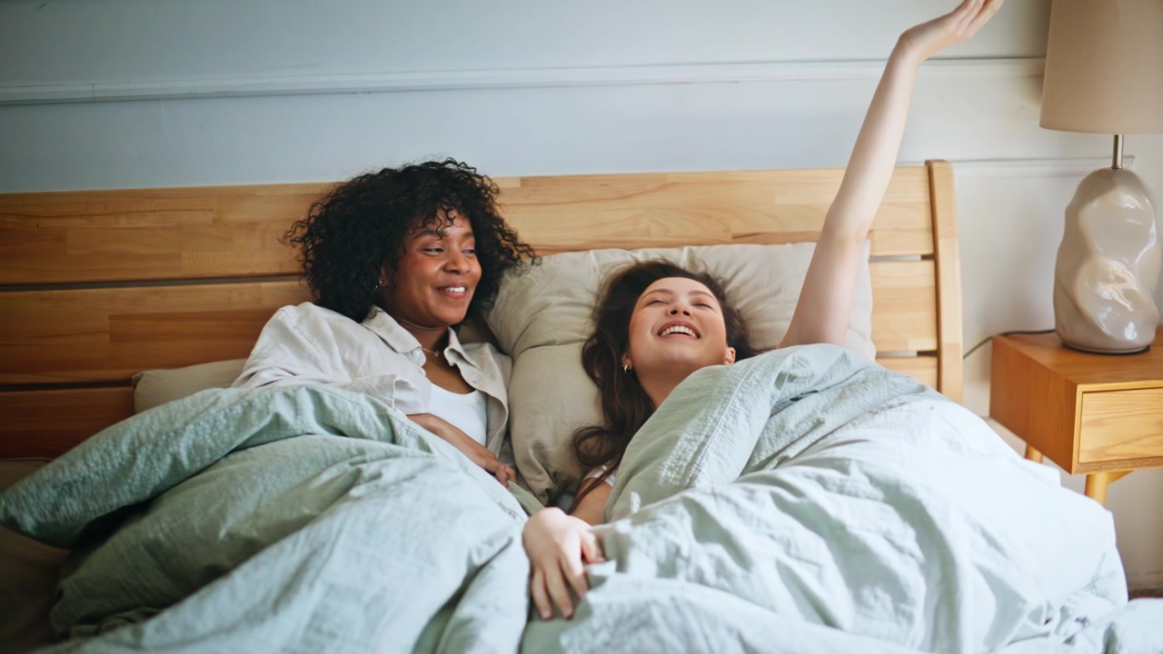 Lesbian couple waking up morning bed. Girlfriends laughing stretching bedroom.
