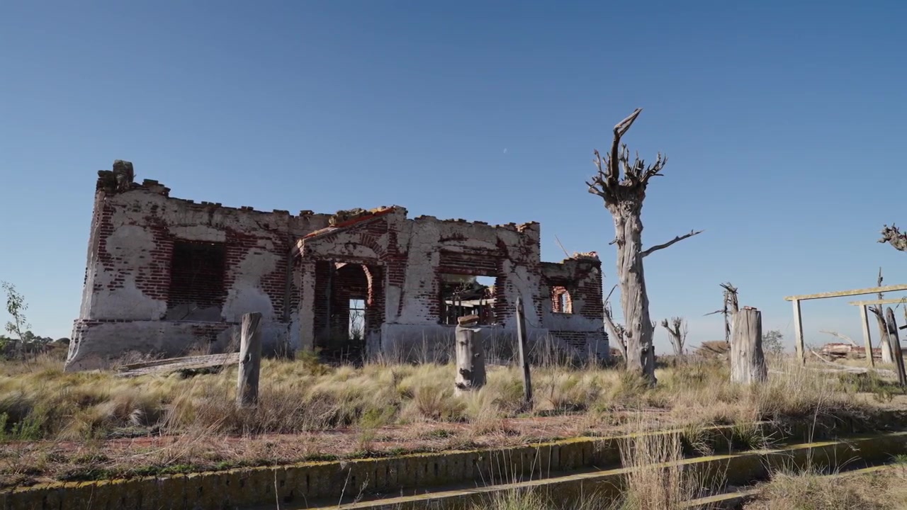 Static right pan of ruined brick building and skeletal dead trees at Villa Epecuén under a vivid blue afternoon sky.