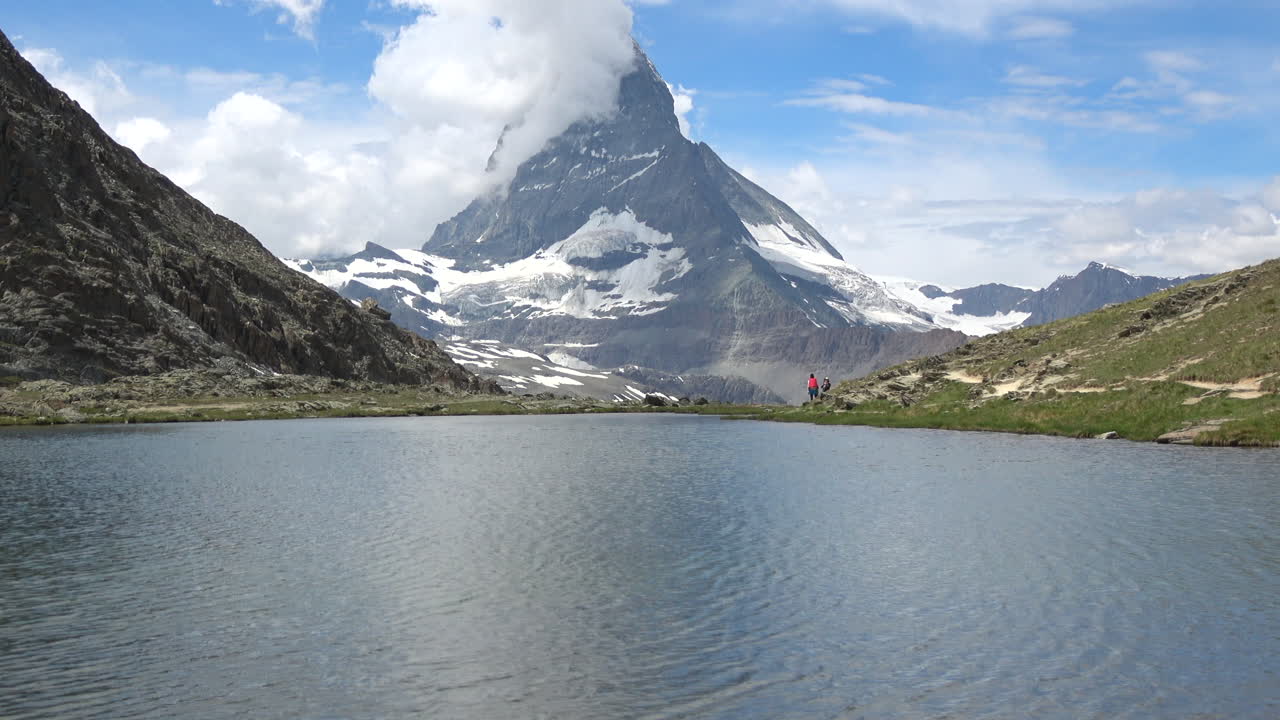 Ampia inquadratura di un calmo lago alpino che riflette l'iconica montagna del Cervino, con escursionisti sulla riva e nuvole che fluttuano sopra, filmato a Zermatt, Vallese, Svizzera
