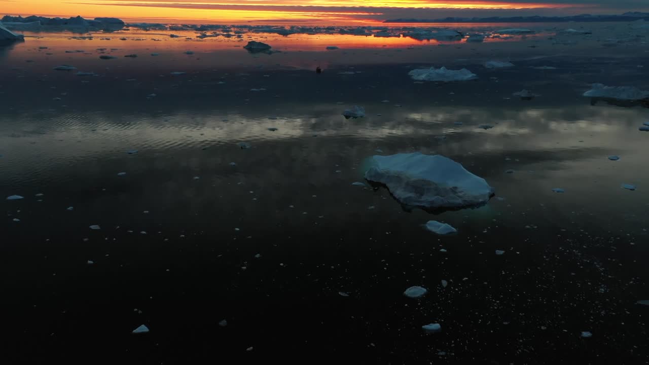 Ilmakuva dronella kuvattuna kelluvista jääkimpaleista tyynellä Jäämerellä eloisan auringonnousun aikana Grönlannin lähellä. Lämmin oranssi valo heijastuu jäiseen veteen dramaattisten pilvien alla.