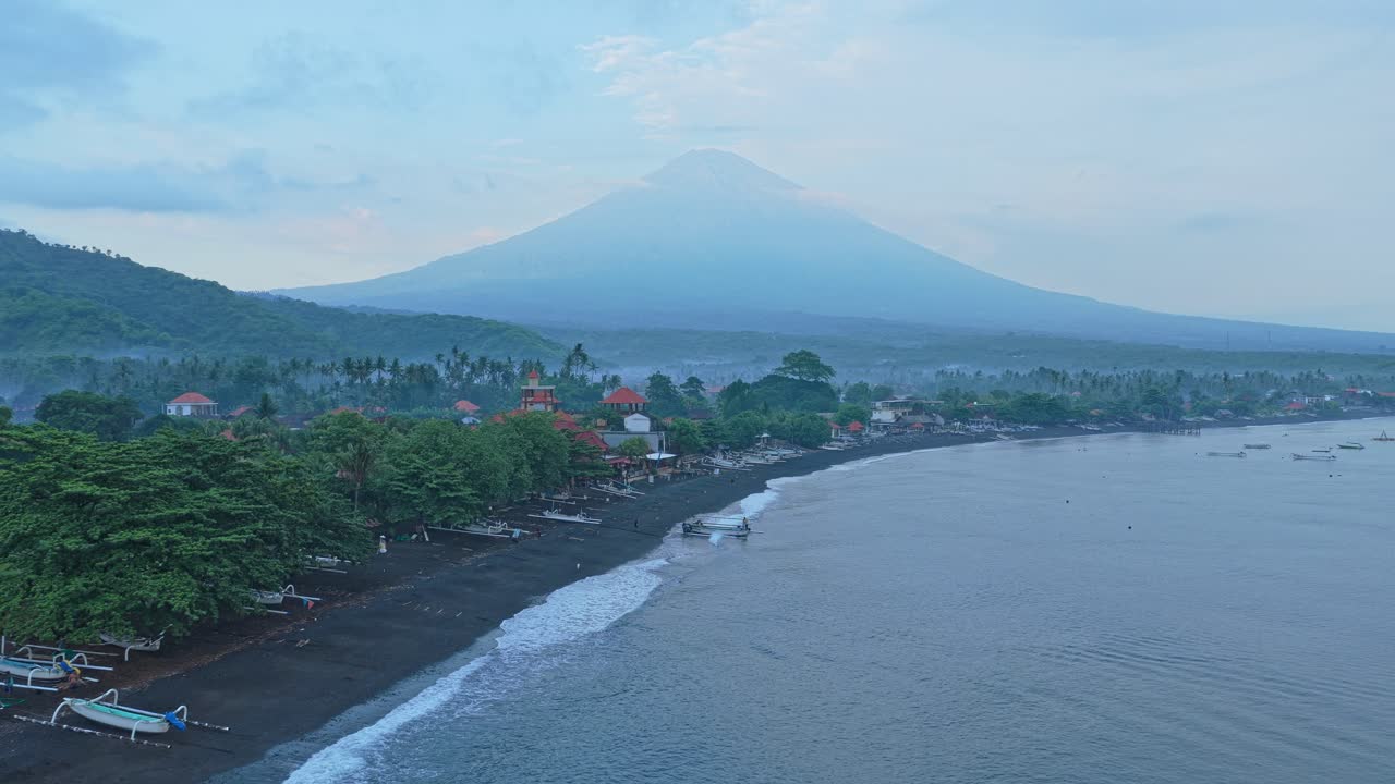 Maestosa vista aerea del villaggio di Amed con la sua caratteristica spiaggia di sabbia nera e le tradizionali barche jukung, sotto l'imponente vulcano Monte Agung.