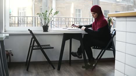 Schönes-Muslimisches-Mädchen-Mit-Hijab-Sucht-Auf-Ihrem-Laptop-Im-Internet-Nach-Etwas-Und-Schreibt-Etwas-Auf,-Während-Sie-Drinnen-An-Einem-Minimalistischen,-Hellen-Ort-Sitzt