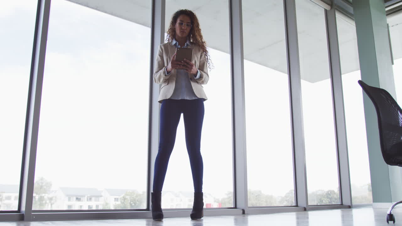 Biracial businesswoman walking and using tablet in modern interiors